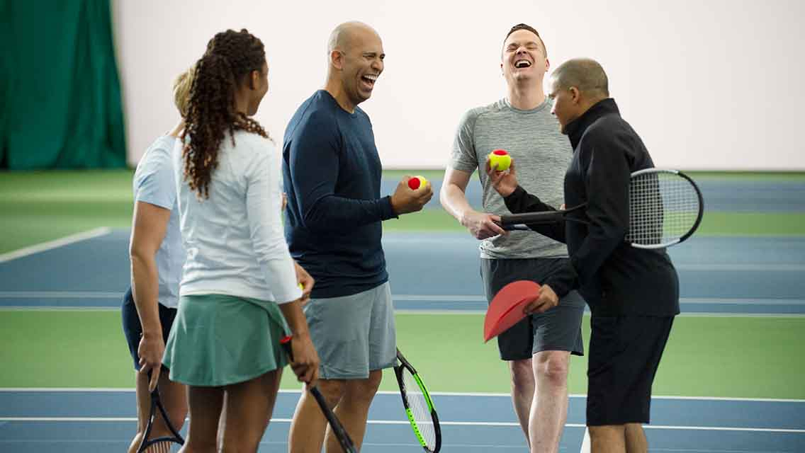 Four friends laughing while holding tennis rackets and balls on a tennis court.