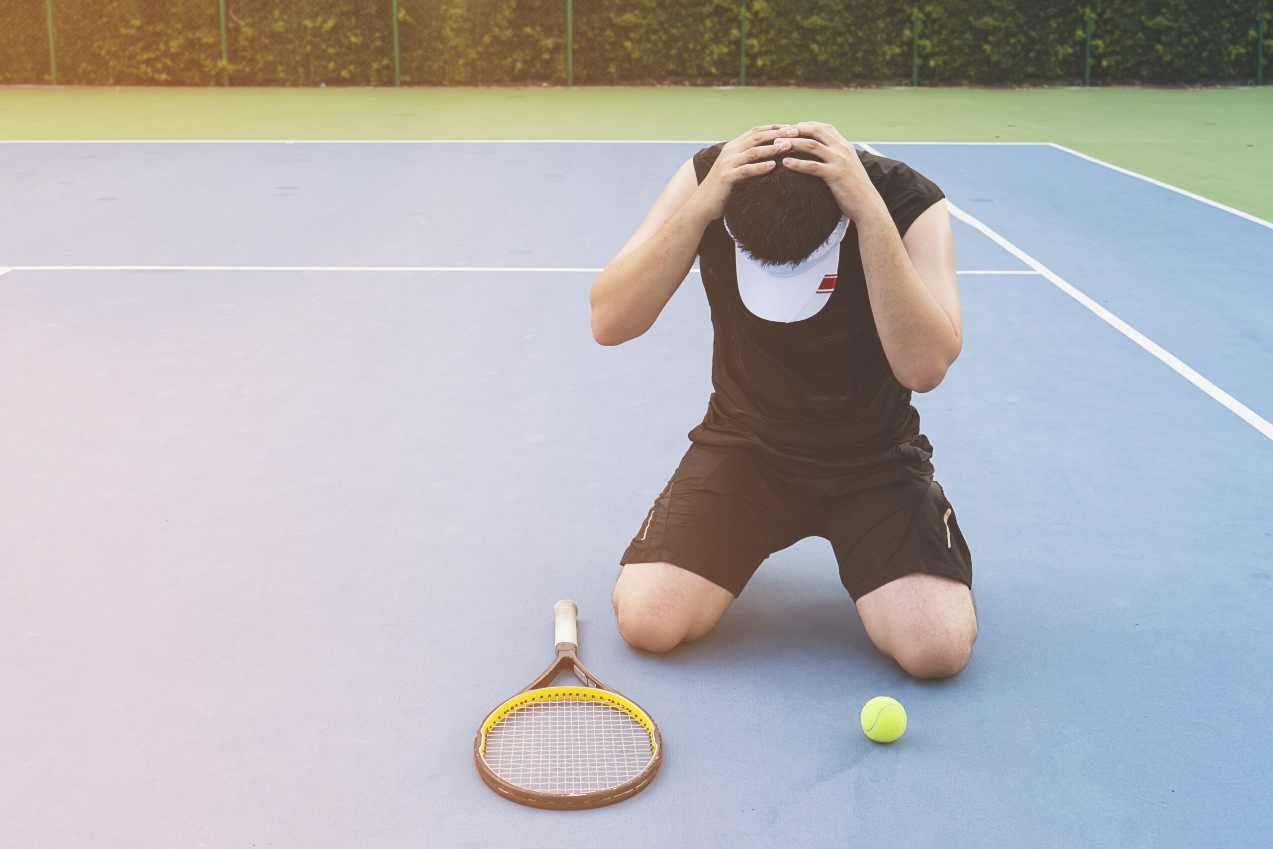 Player kneeling on court, holding head in frustration beside tennis racket and ball.